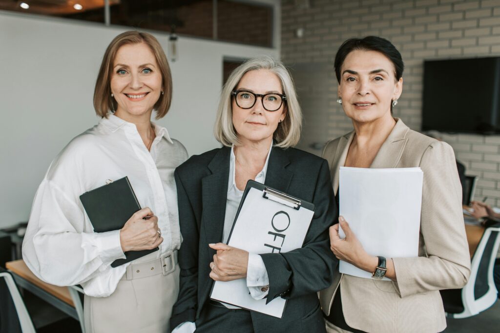 Three confident businesswomen in professional attire holding papers in an office setting.